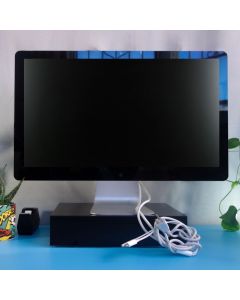 A good as new Thunderbolt Display Monitor 27'' sits unpluged on a black wooden shape. Along the side there are a cactus with comic book style decoration on its medium sized planter next to an office black tape dispenser, on top a sky blue desk. In the bac