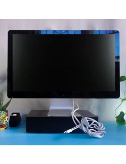 A good as new Thunderbolt Display Monitor 27'' sits unpluged on a black wooden shape. Along the side there are a cactus with comic book style decoration on its medium sized planter next to an office black tape dispenser, on top a sky blue desk. In the bac